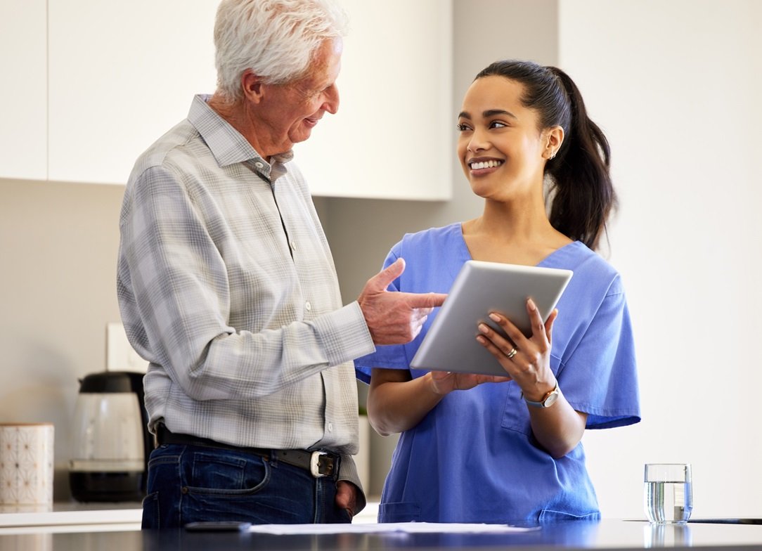 A smiling female caregiver in blue scrubs leaning toward an elderly man in a checkered shirt who is being served a healthy meal tray.